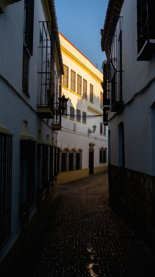 Free A picturesque narrow street in Ronda, Spain, highlighted by morning sunlight casting shadows. Stock Photo