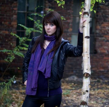 Stylish young woman in leather jacket and scarf posing outdoors beside a tree.