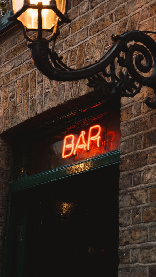 Free Neon bar sign glowing on a brick building facade under a classic streetlamp at night. Stock Photo