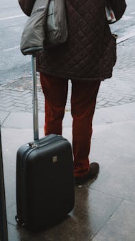Back view of a person standing with luggage on a wet urban sidewalk.
