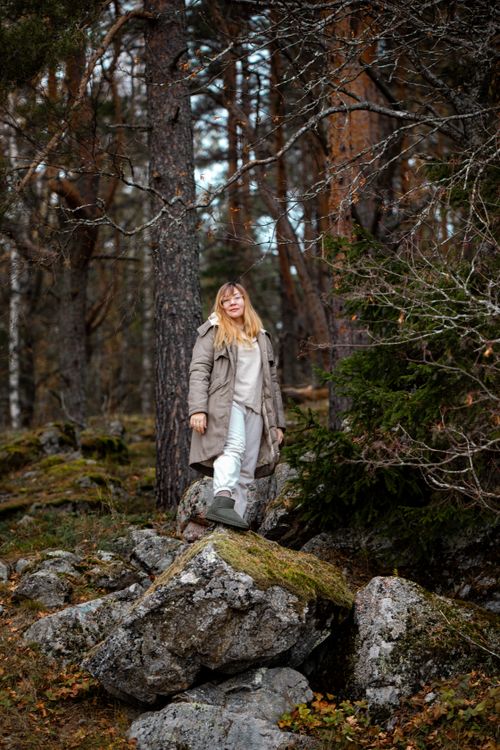 Free Woman in winter jacket standing on rocks in a serene forest scene. Stock Photo