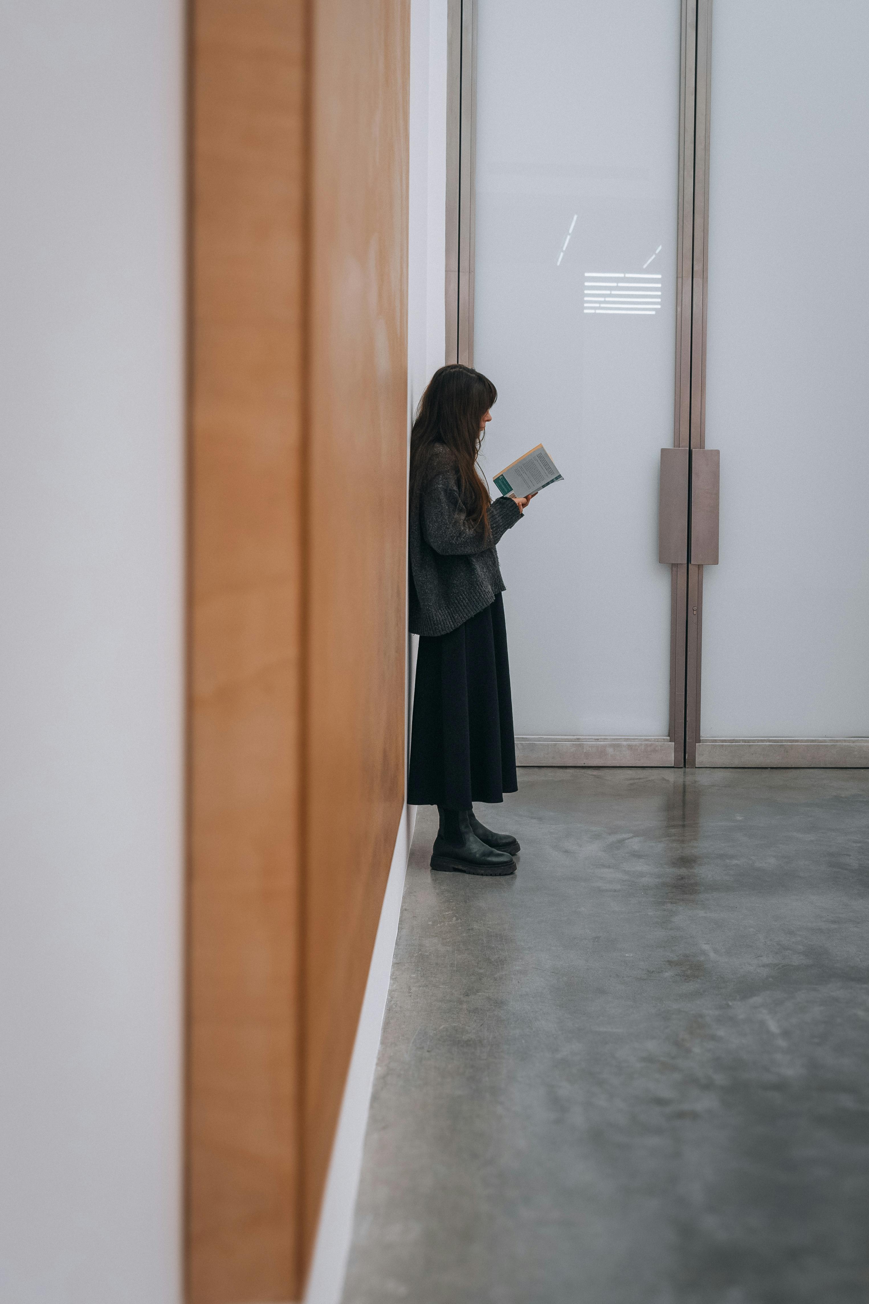 A woman leans against a wall reading a book in a modern indoor hallway in London, England.