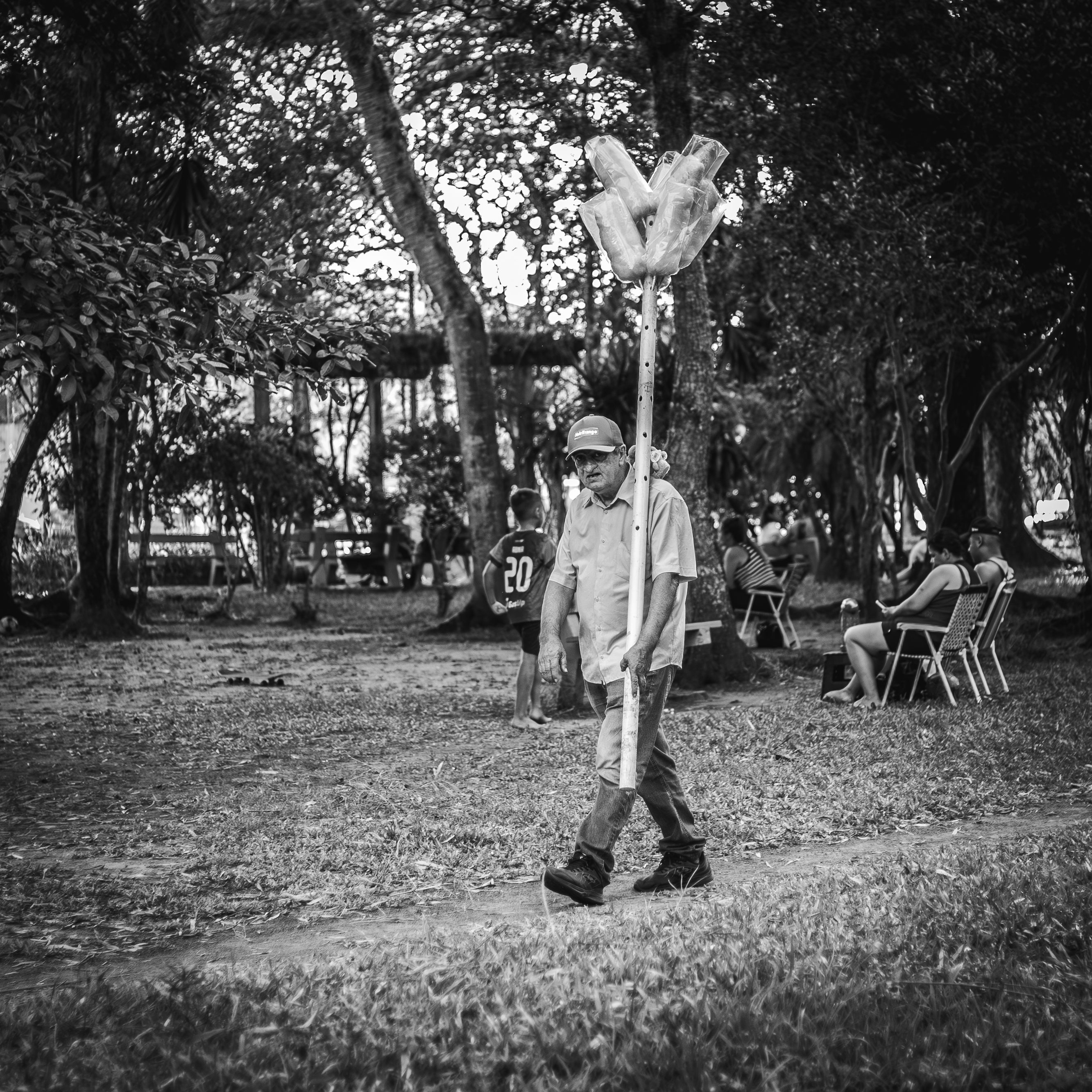Elderly man with cap walking through park holding a large object, black and white.