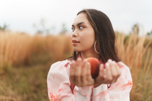Woman Holding a Fruit Standing on a Field and Looking Away 