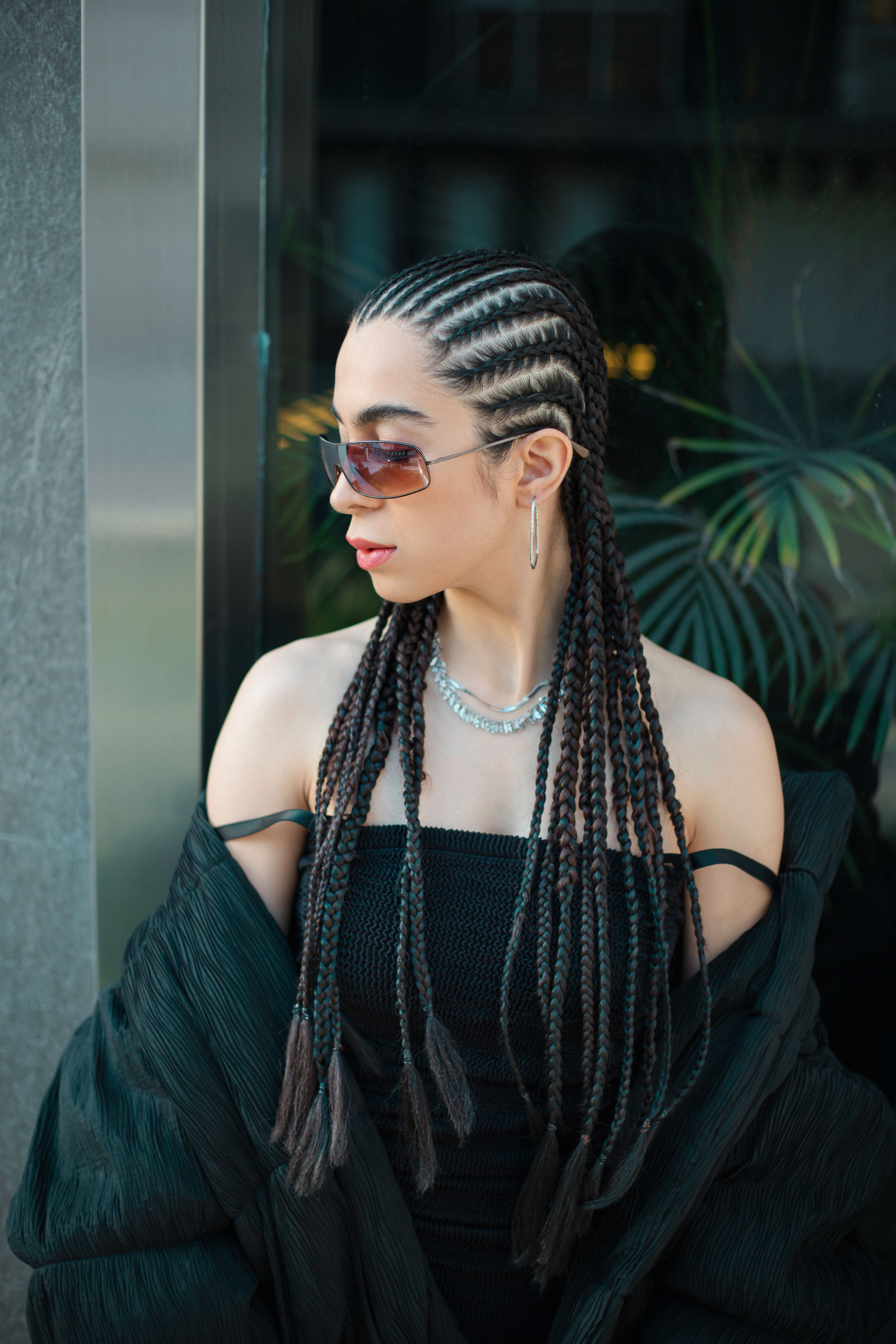 Fashionable woman with braided hair and sunglasses posing outdoors with plants in the background.