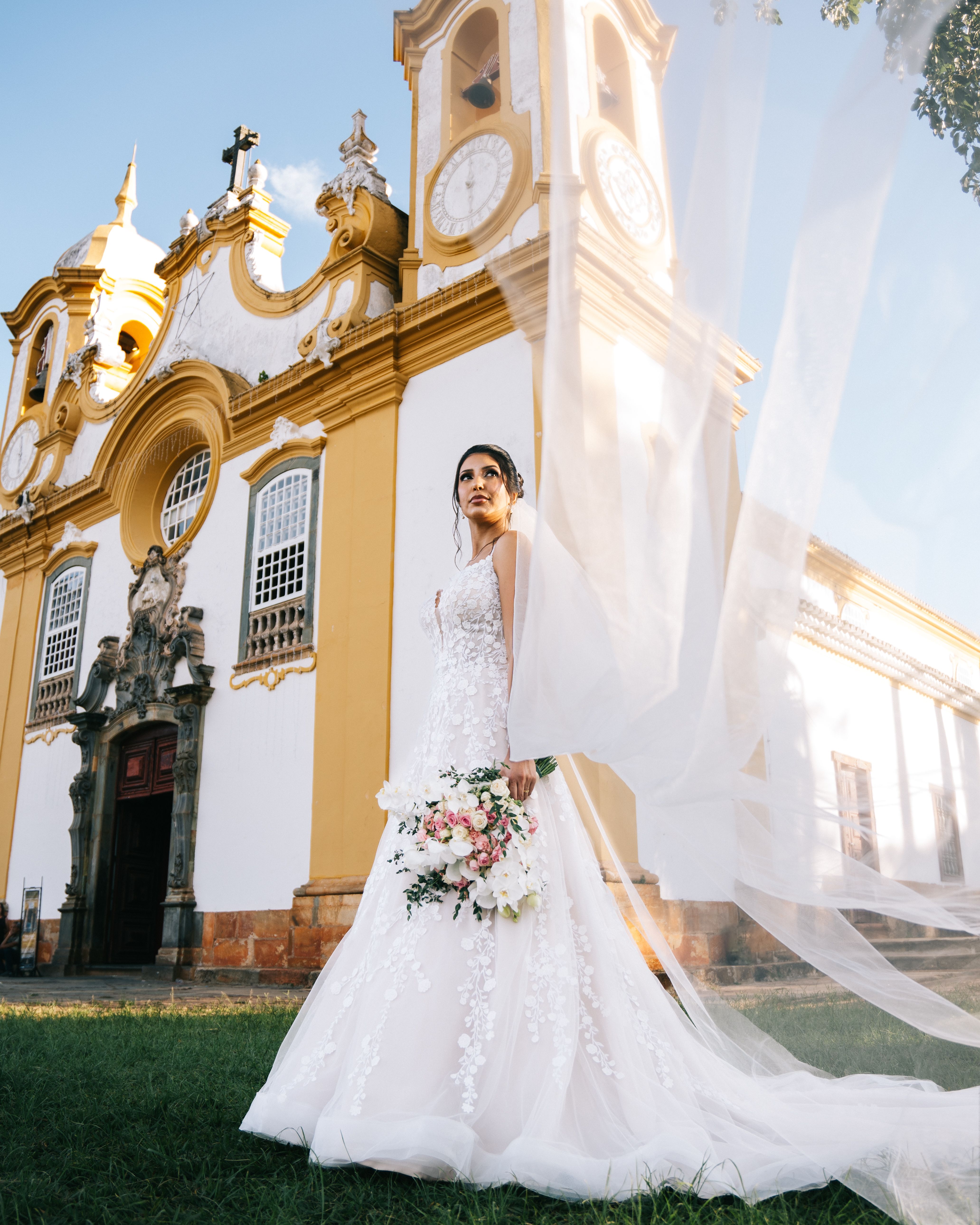 Elegant bride in wedding dress standing before a historic church in Tiradentes, MG, Brazil, holding a bouquet.