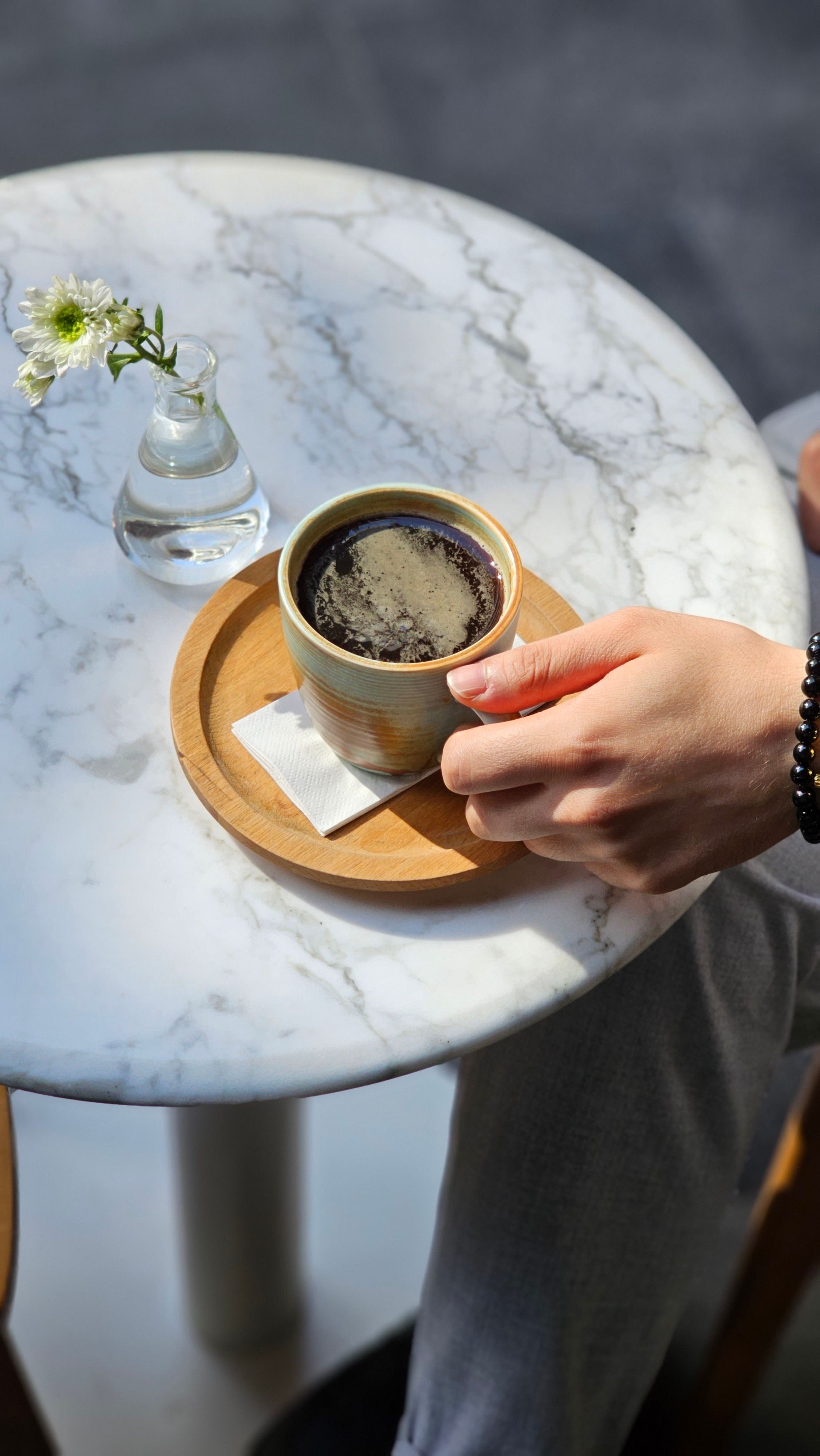 A hand holding a cup of coffee on a marble table with a flower vase next to it.