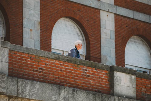 An older man is standing on a ledge looking out over a brick wall