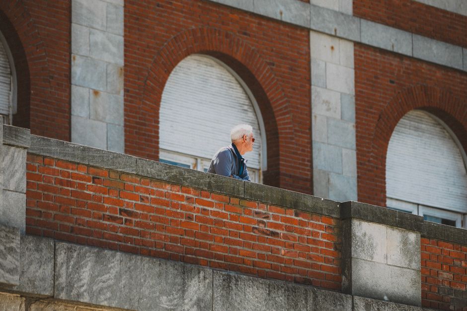 Hombre mayor con cabello gris mirando desde un balcón de ladrillo en la ciudad.