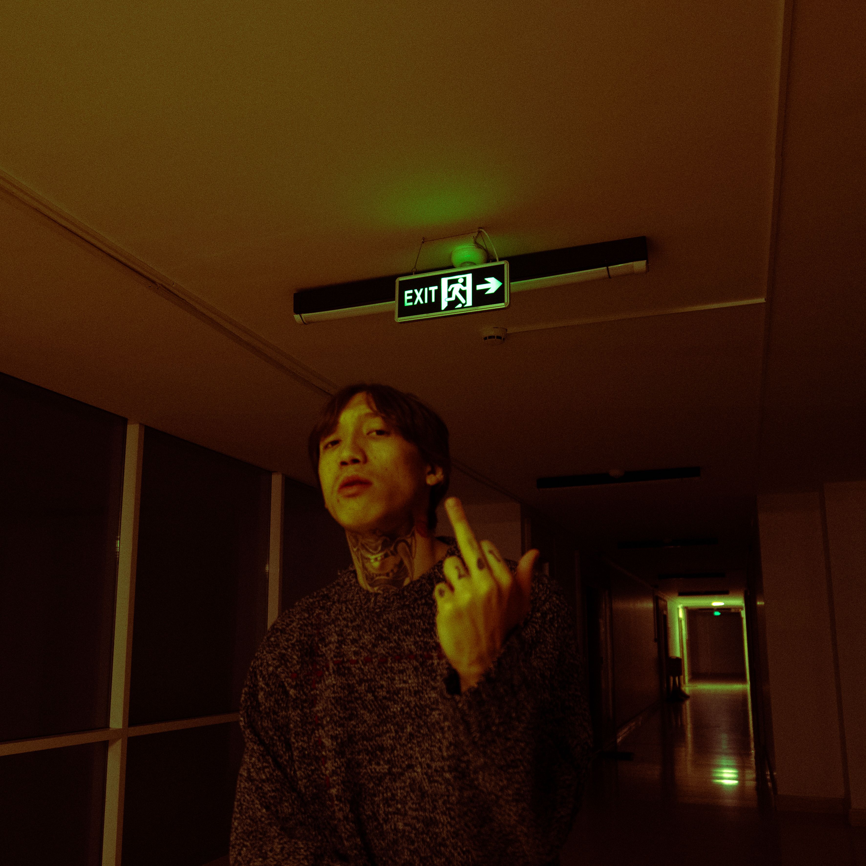 A man with tattoos stands under an exit sign in a dimly lit corridor at night.