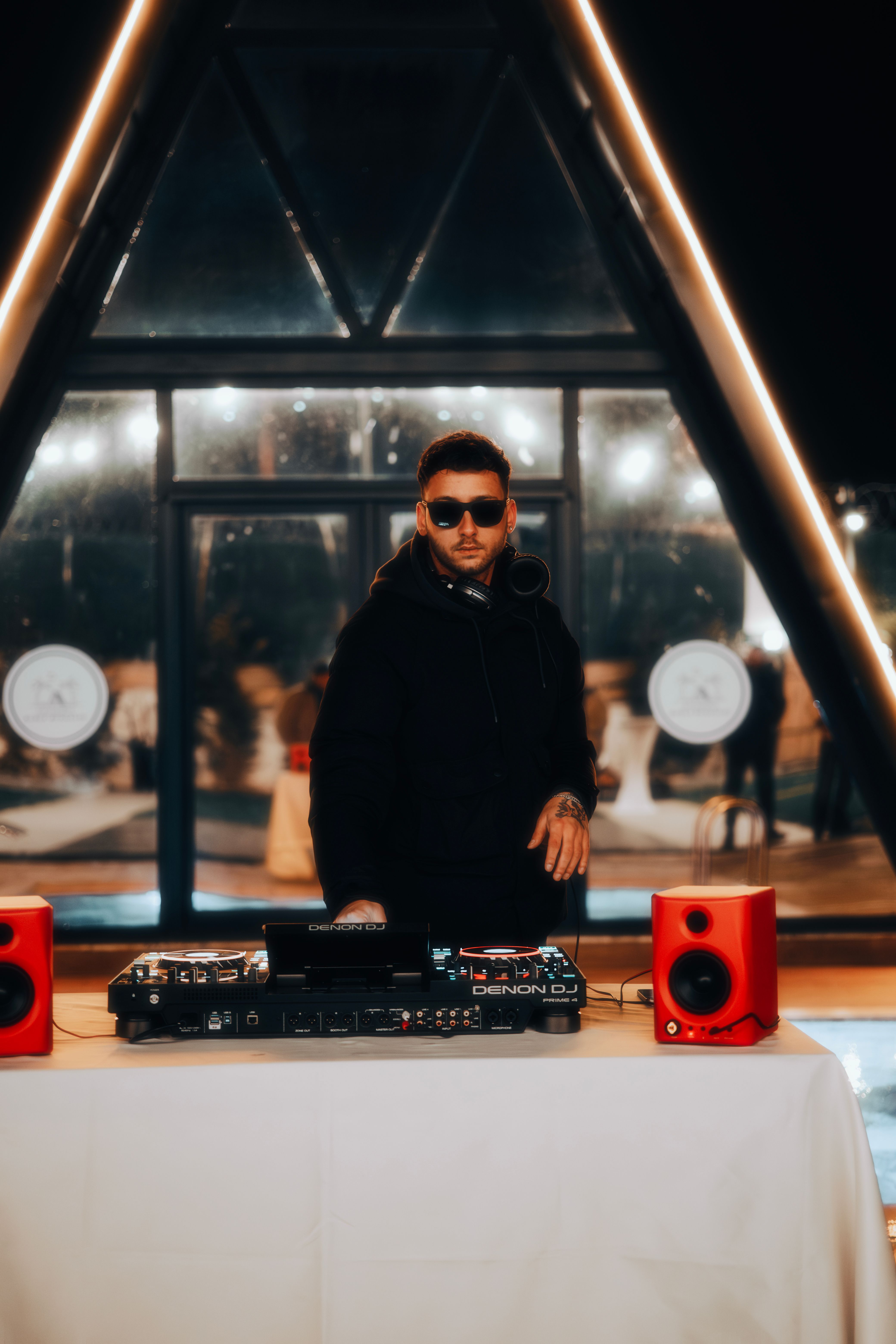 DJ with sunglasses performs indoors at night, using console and red speakers.