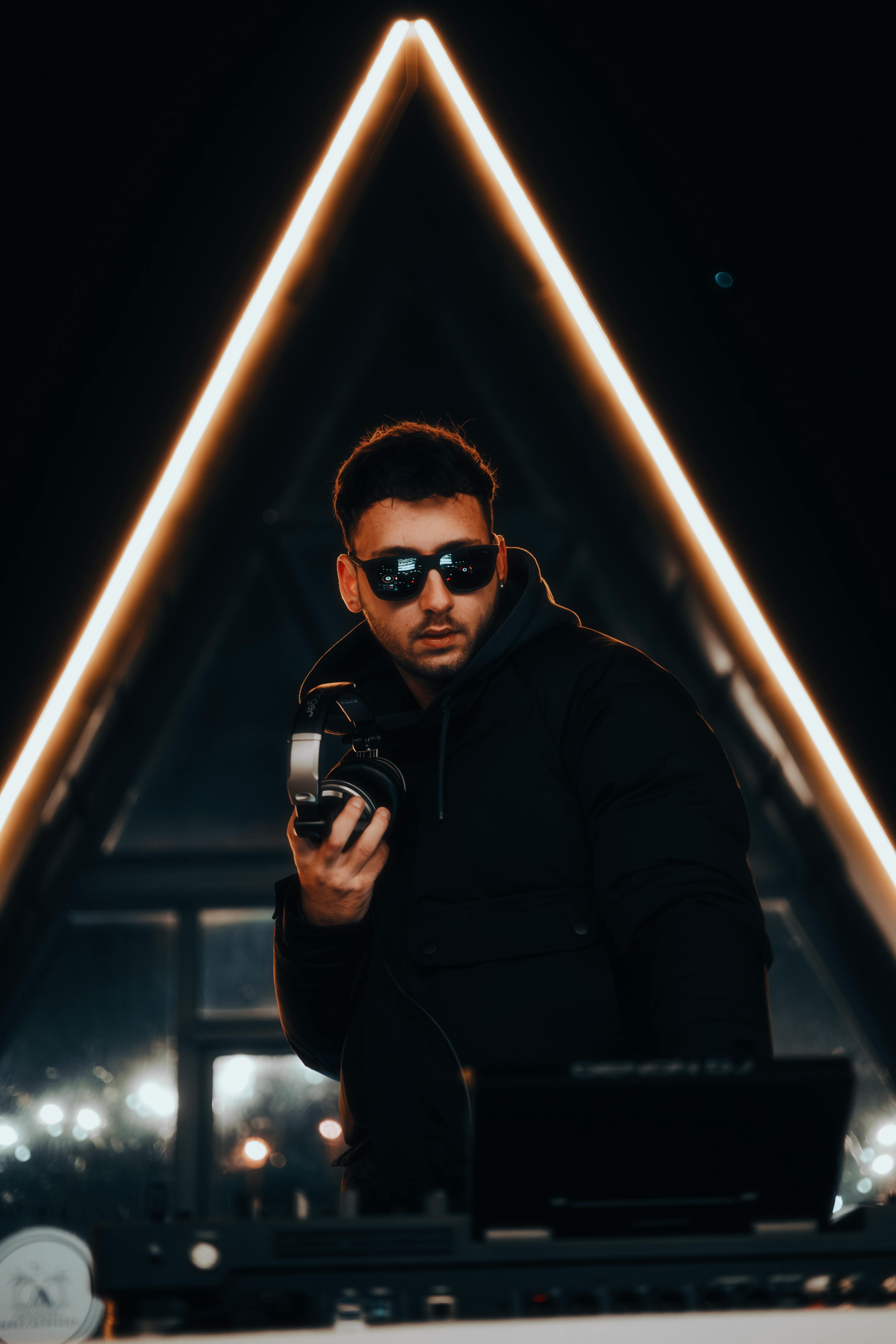 DJ in sunglasses holding headphones, performing live at a vibrant night event with triangular neon lights.