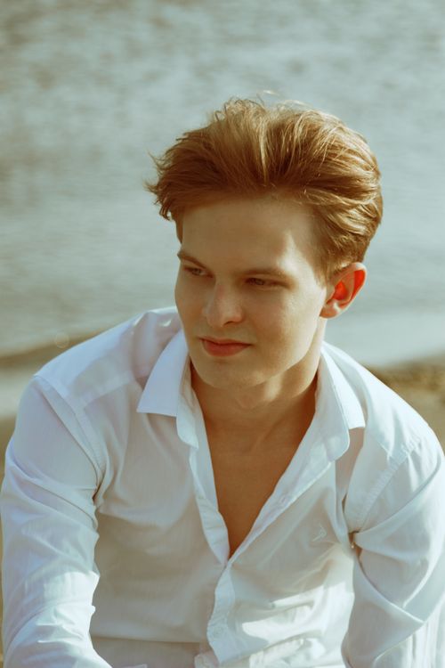 Free Young man in a white shirt sitting by the water, captured in natural sunlight. Stock Photo