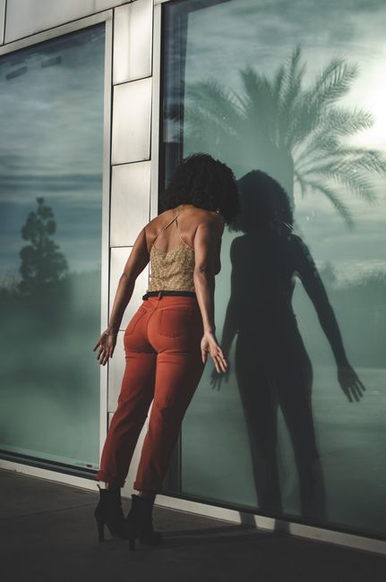 Stylish woman in urban setting leaning against glass wall reflecting a palm tree silhouette.