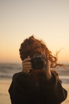 A redheaded woman with tousled hair uses a camera by the sea at sunset.