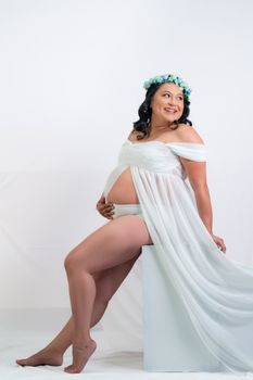 Pregnant woman in white dress and floral crown smiling against a white background.