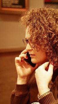 Woman with curly hair and eyeglasses talking on a smartphone indoors.