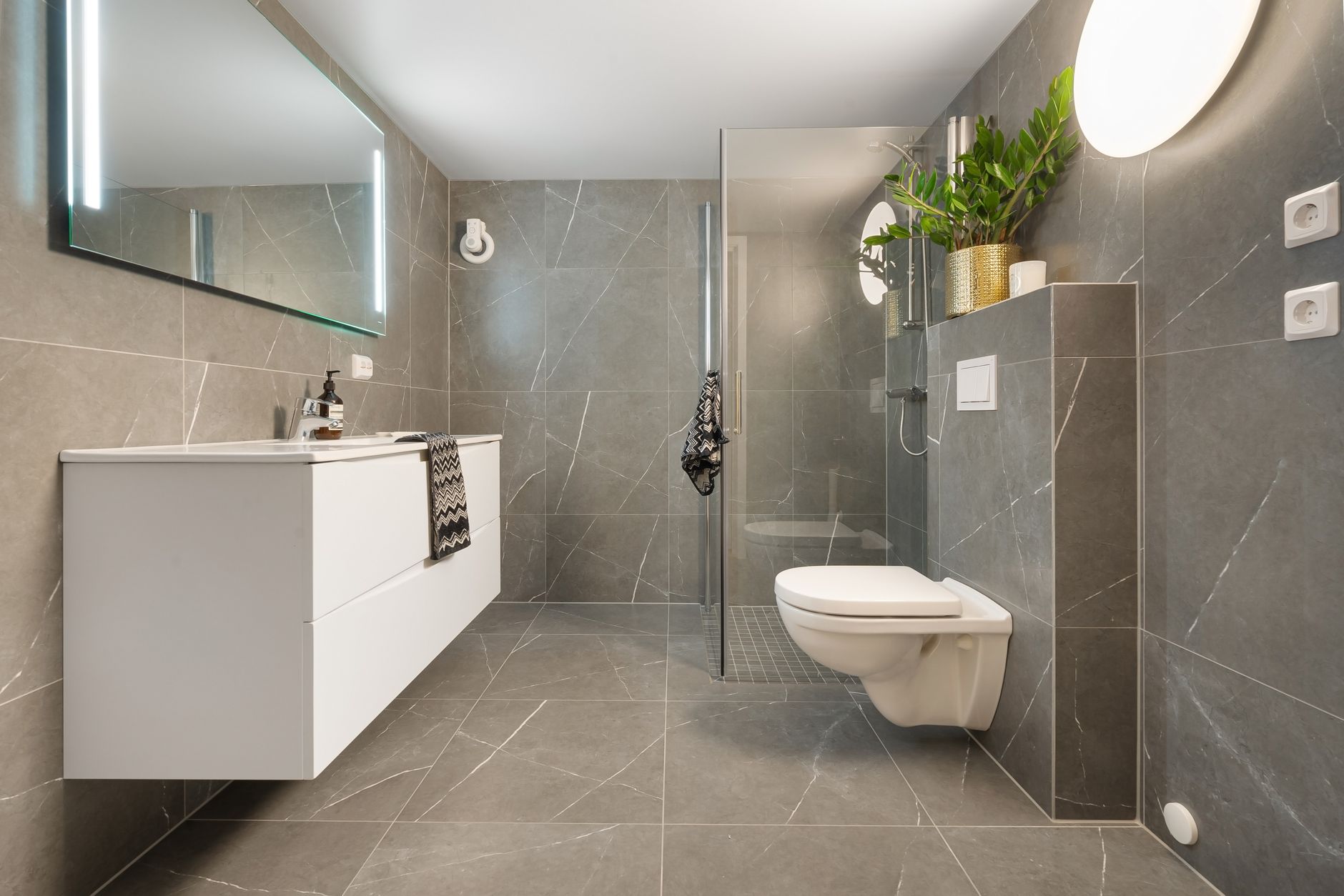 Stylish gray tiled bathroom featuring a shower, toilet, and illuminated mirror.