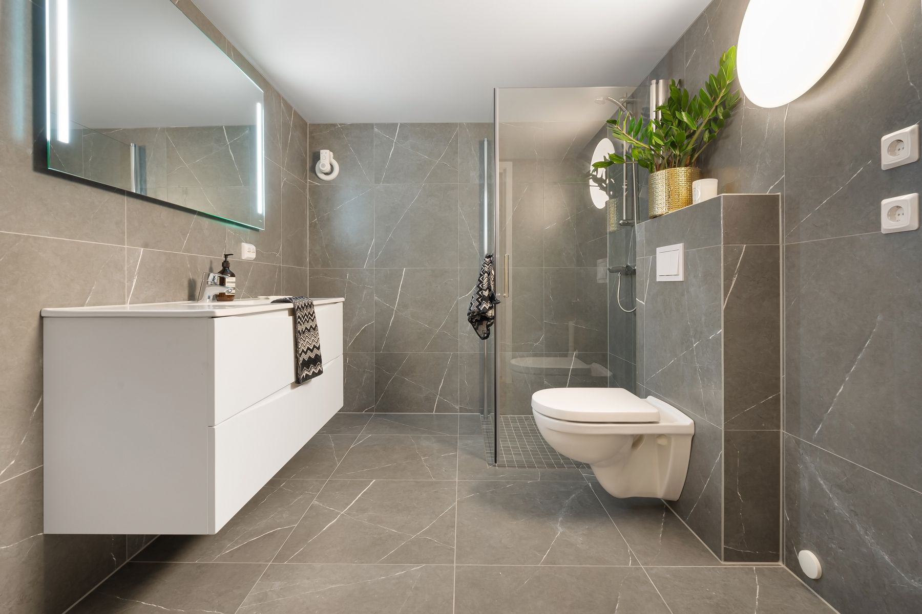 Modern grey tiled bathroom with glass shower, toilet and illuminated mirror