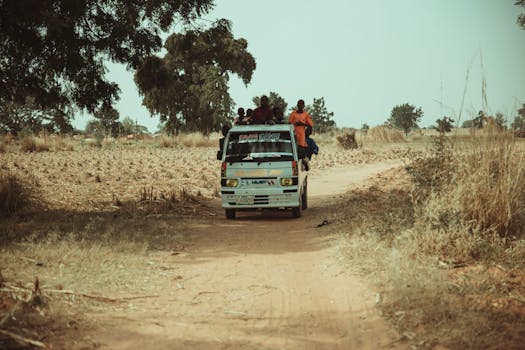 A group of people riding on a van through a rural dirt road surrounded by fields and trees.