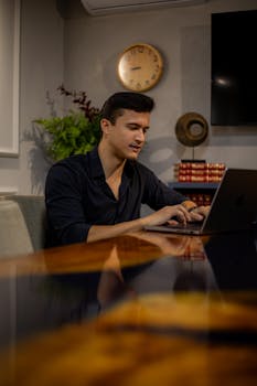 Caucasian man focused on work at laptop, seated at a modern office table with books and decor.