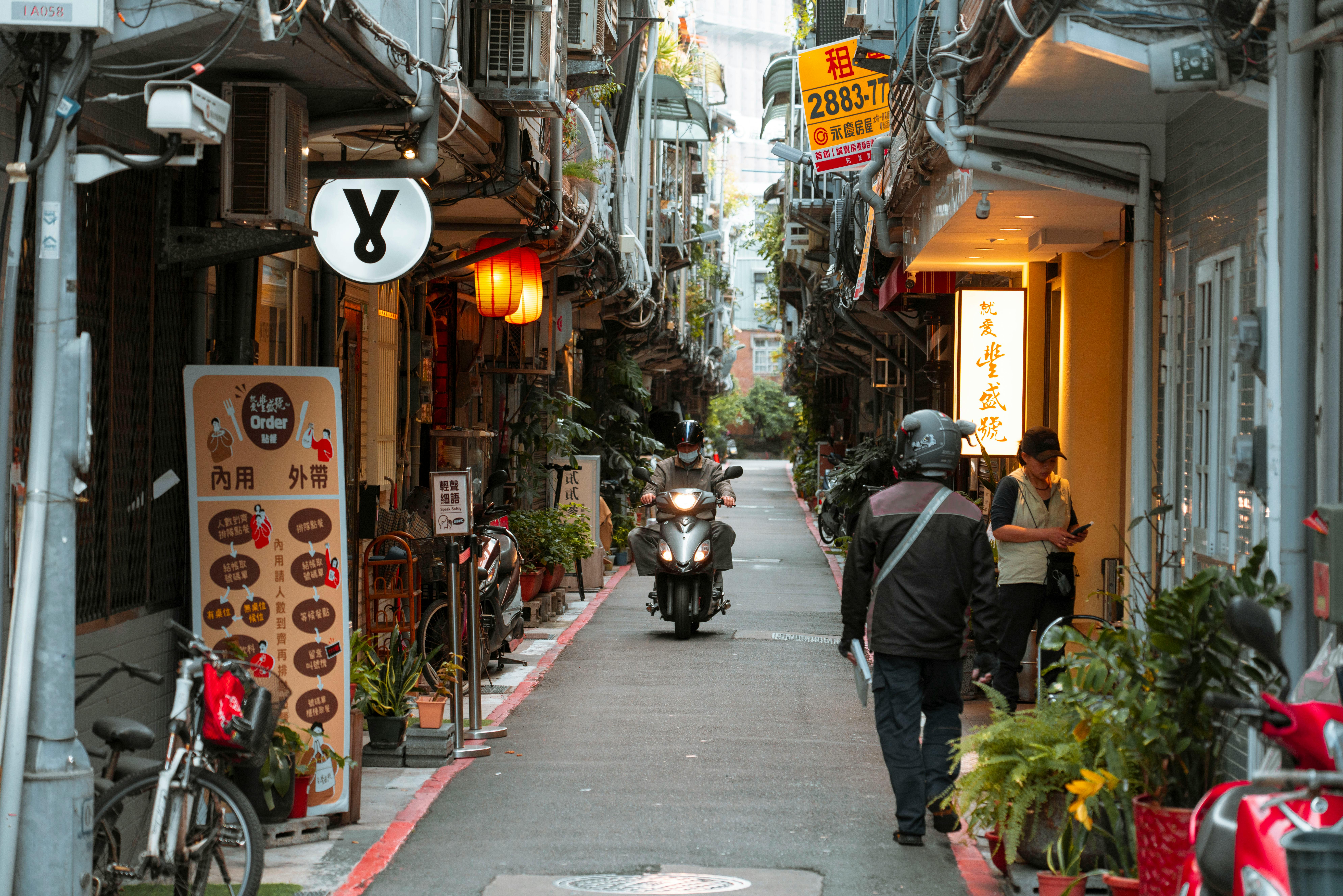 A bustling Asian alley with scooters, eateries, and vibrant city life during day time.