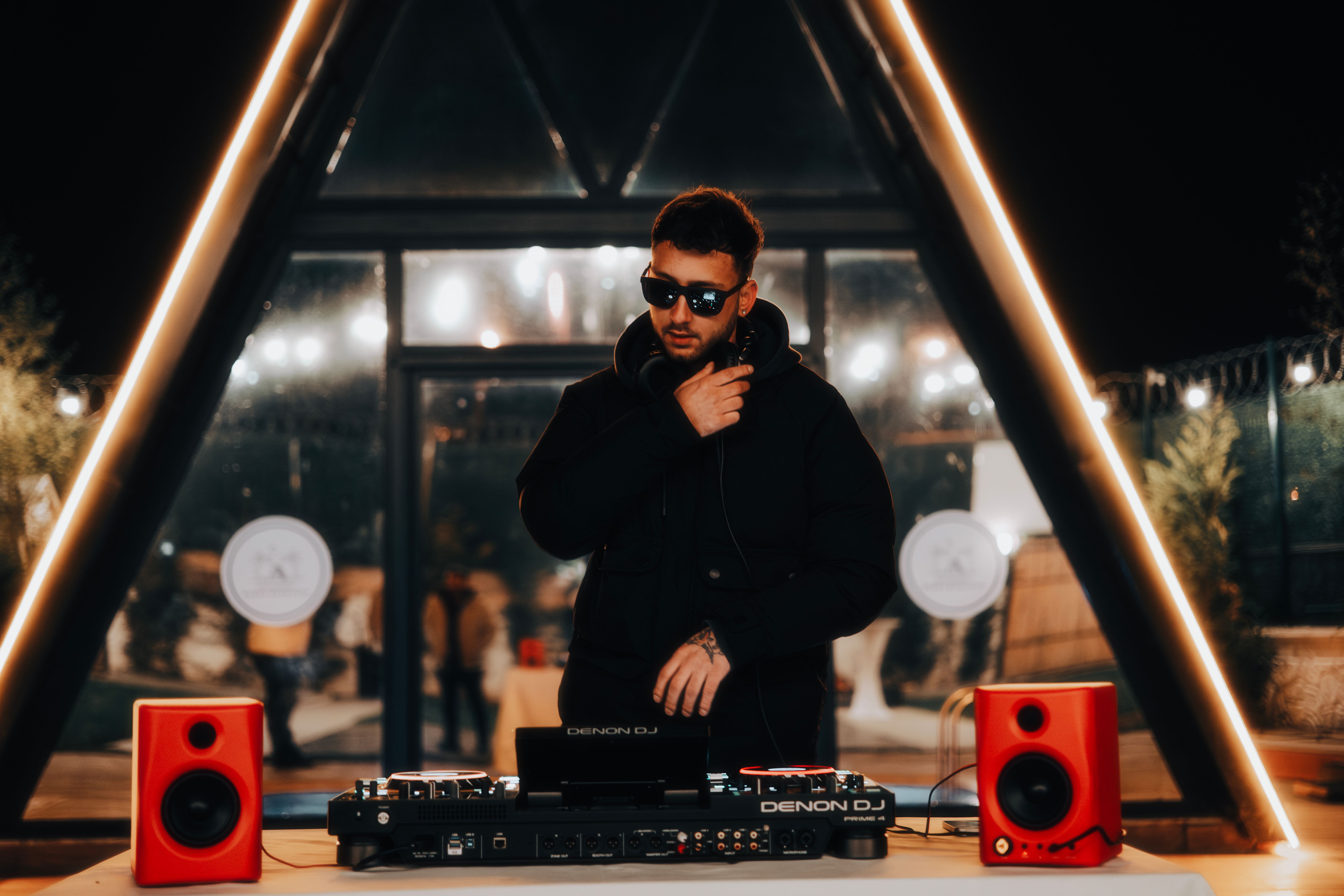 DJ with sunglasses mixing music outdoors at night, surrounded by neon lights and electronic equipment.