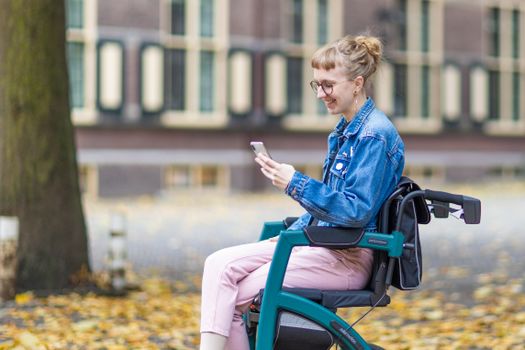 Smiling woman in wheelchair using smartphone in an autumn park in Amsterdam.
