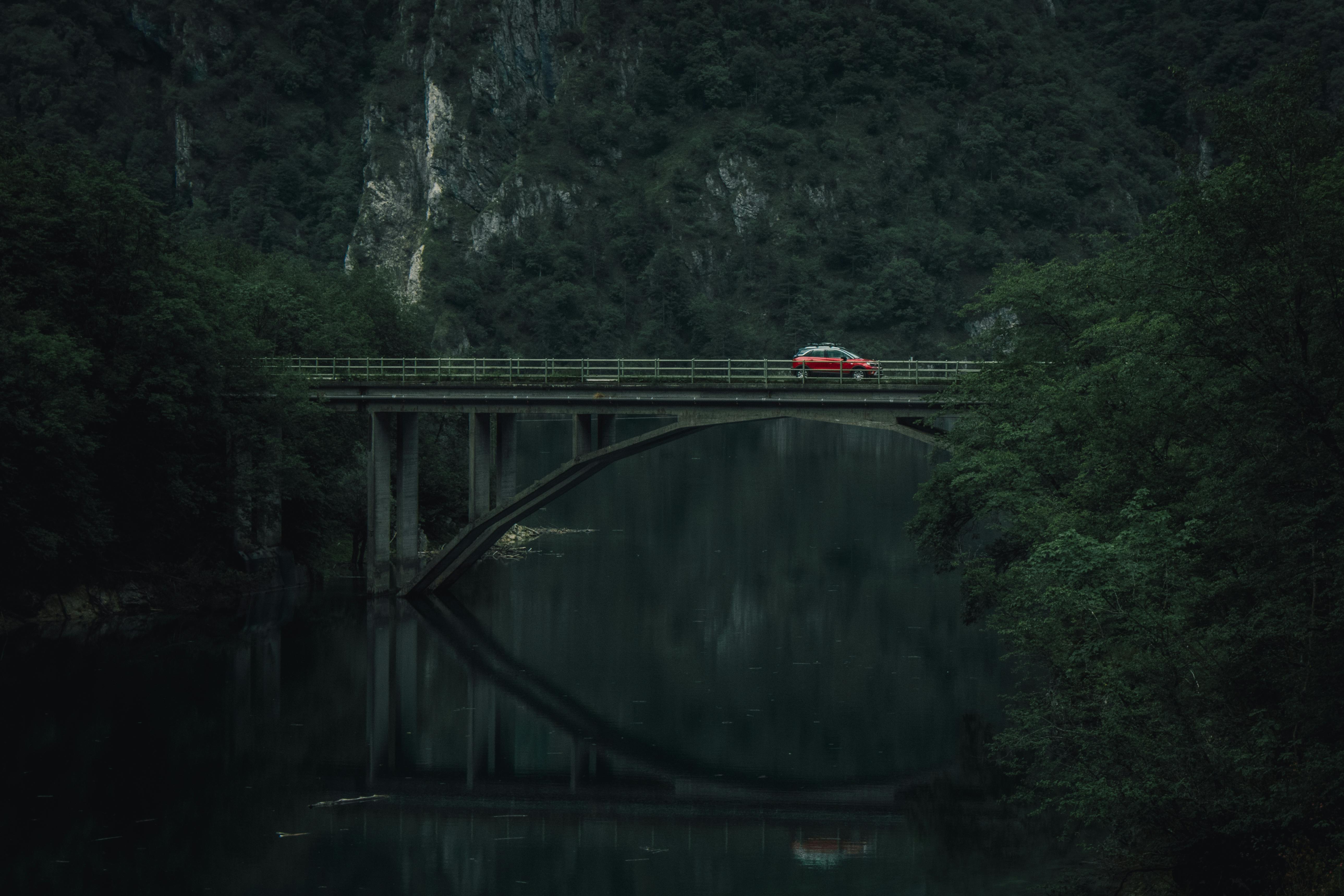 A lone red car crosses a bridge over a calm river in a forest setting with reflections and muted tones.