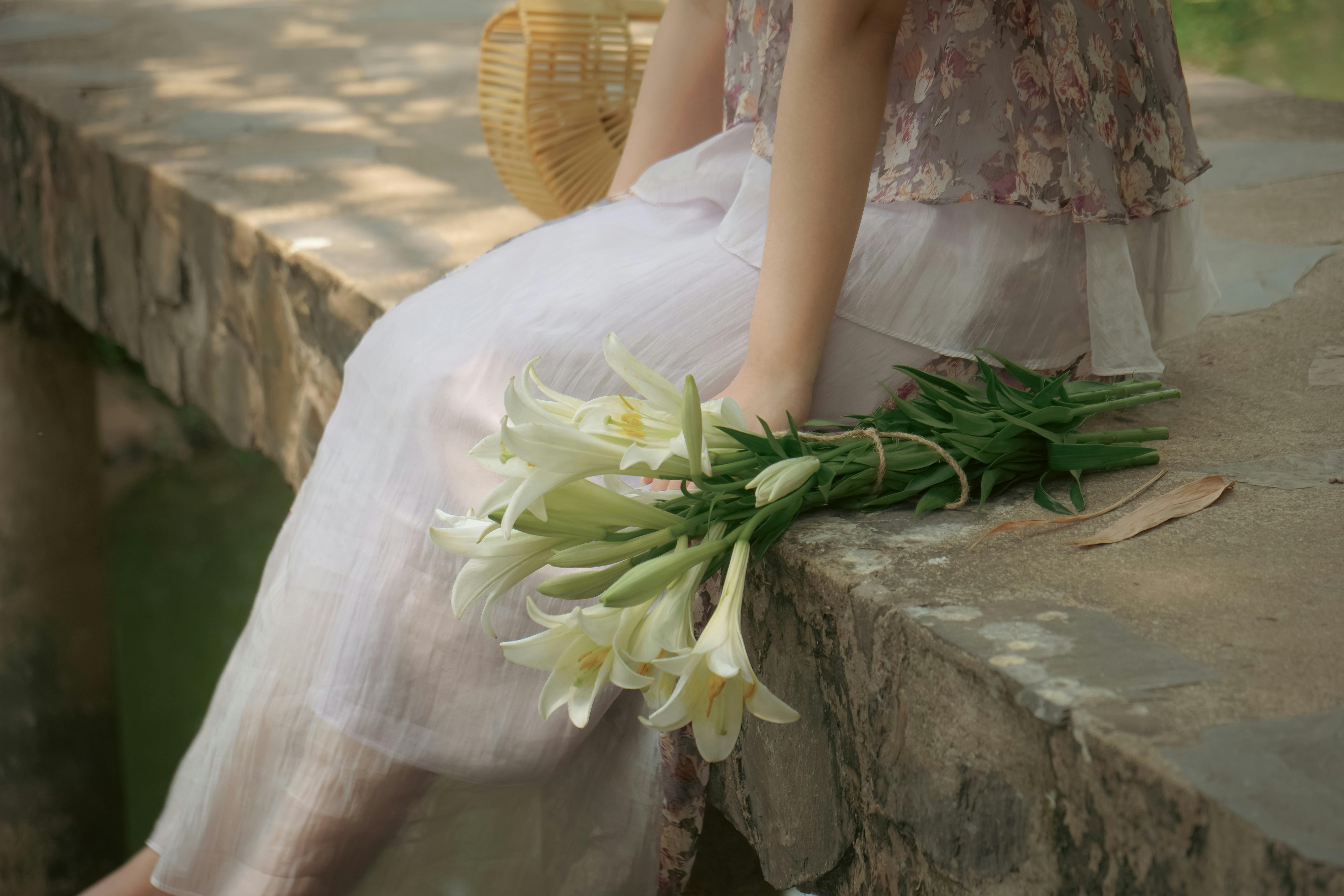 A woman in a white dress holding a bouquet of white lilies on a stone ledge outdoors in daylight.