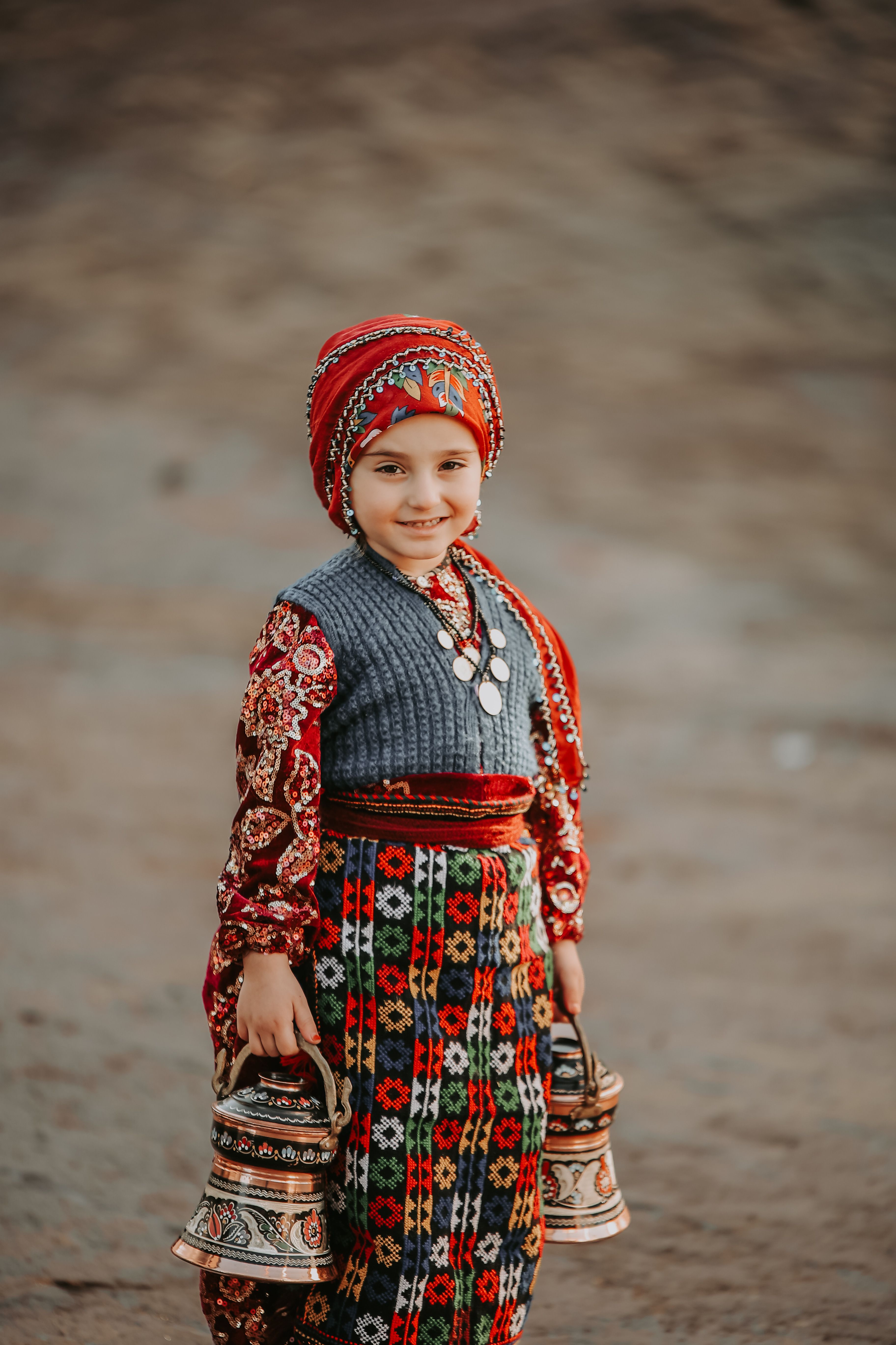Smiling girl in colorful traditional Turkish clothing holding decorative jugs outdoors.
