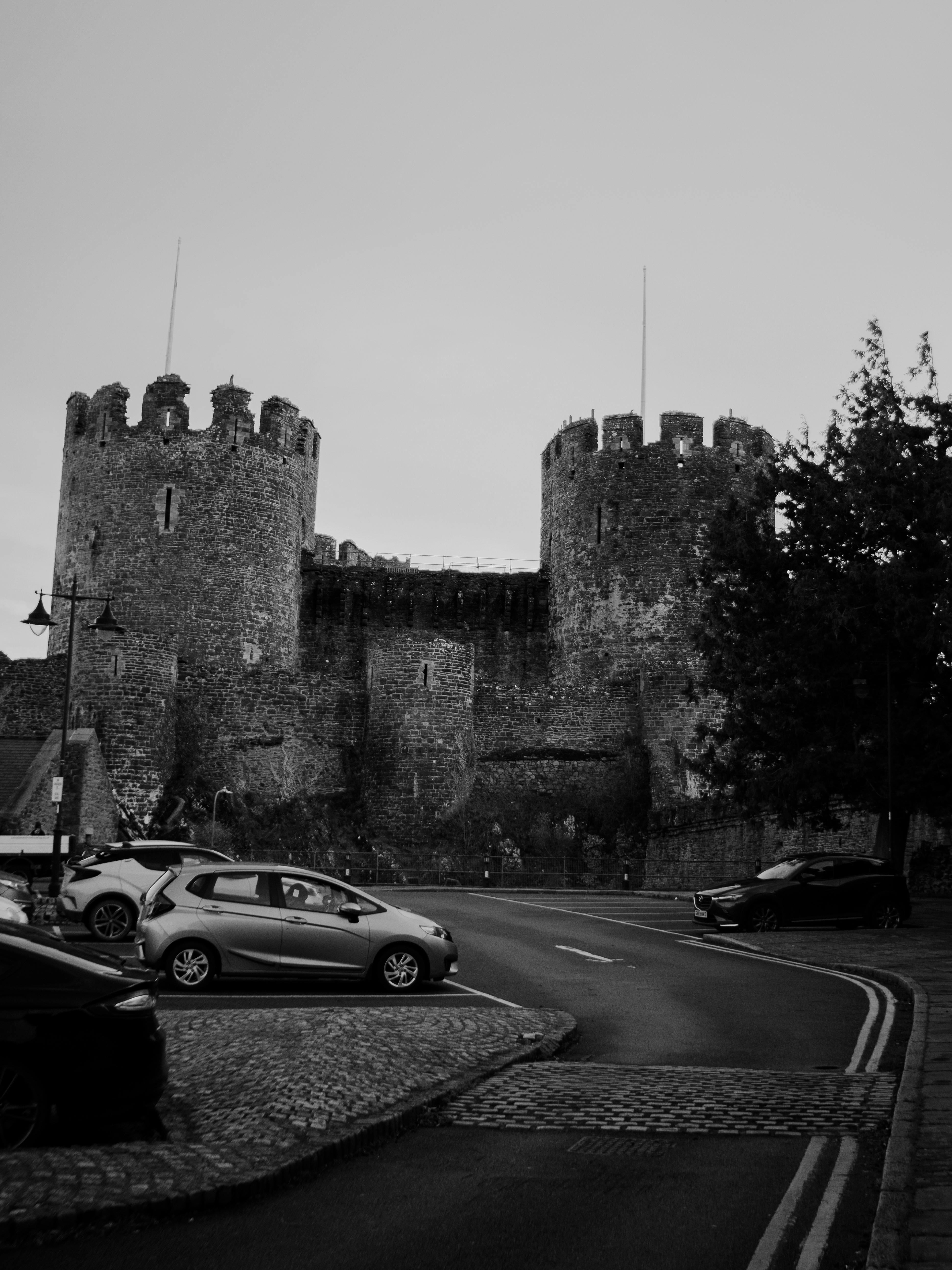 Black and white photo of Conwy Castle with nearby streets and parked cars in Wales, UK.