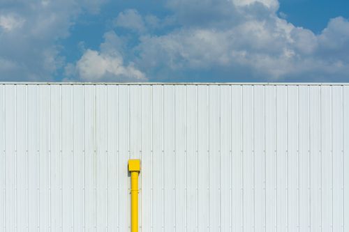 Free A minimalist image of a sunlit white wall featuring a vibrant yellow pipe under a blue sky. Stock Photo