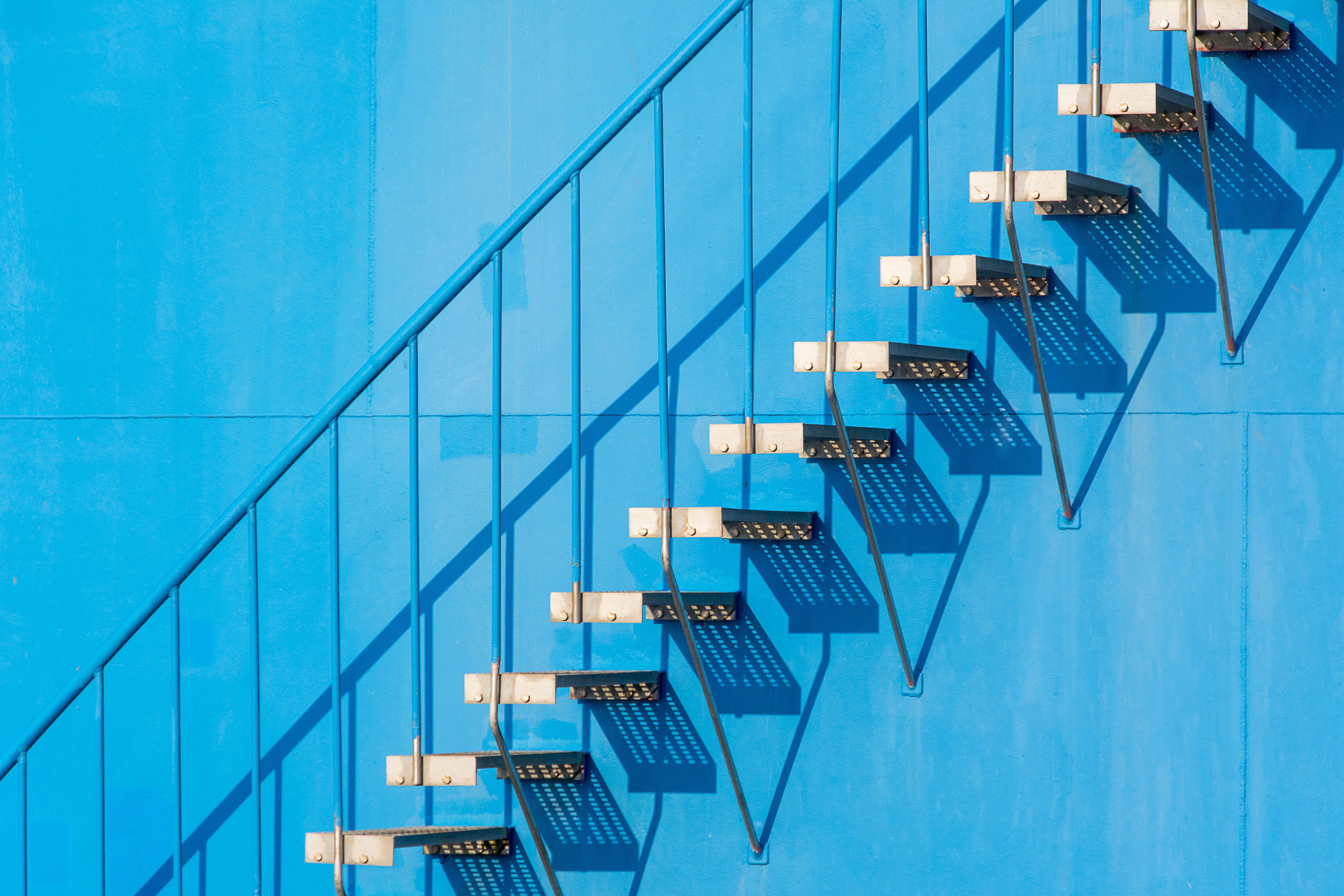 A minimalist view of a steel staircase casting shadows on a blue wall in sunlight.