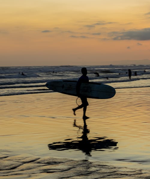 Silhouette of a Person Holding a Surfboard Running on a Beach at Sunset