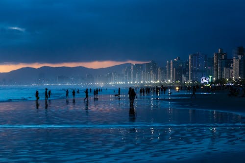 Silhouettes of People on the Ocean Coast with View of Illuminated Waterfront Skyscrapers and Mountains in the Background 