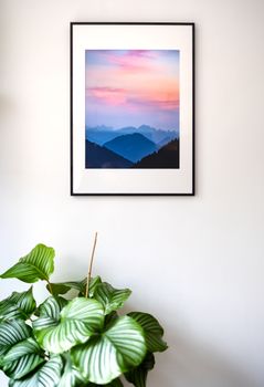 A framed mountain landscape photo hanging on a white wall above a potted plant.