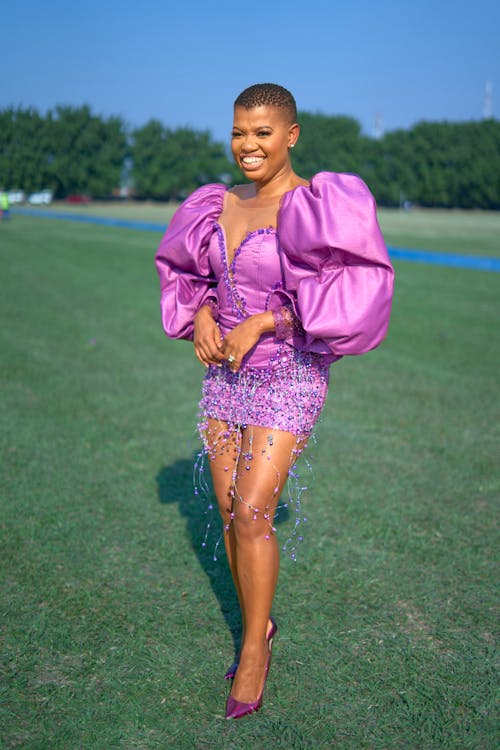 Woman in a Purple Dress and Heels Standing on a Meadow and Smiling 