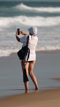 A woman captures ocean waves on a beach with her smartphone, enjoying a sunny summer day.