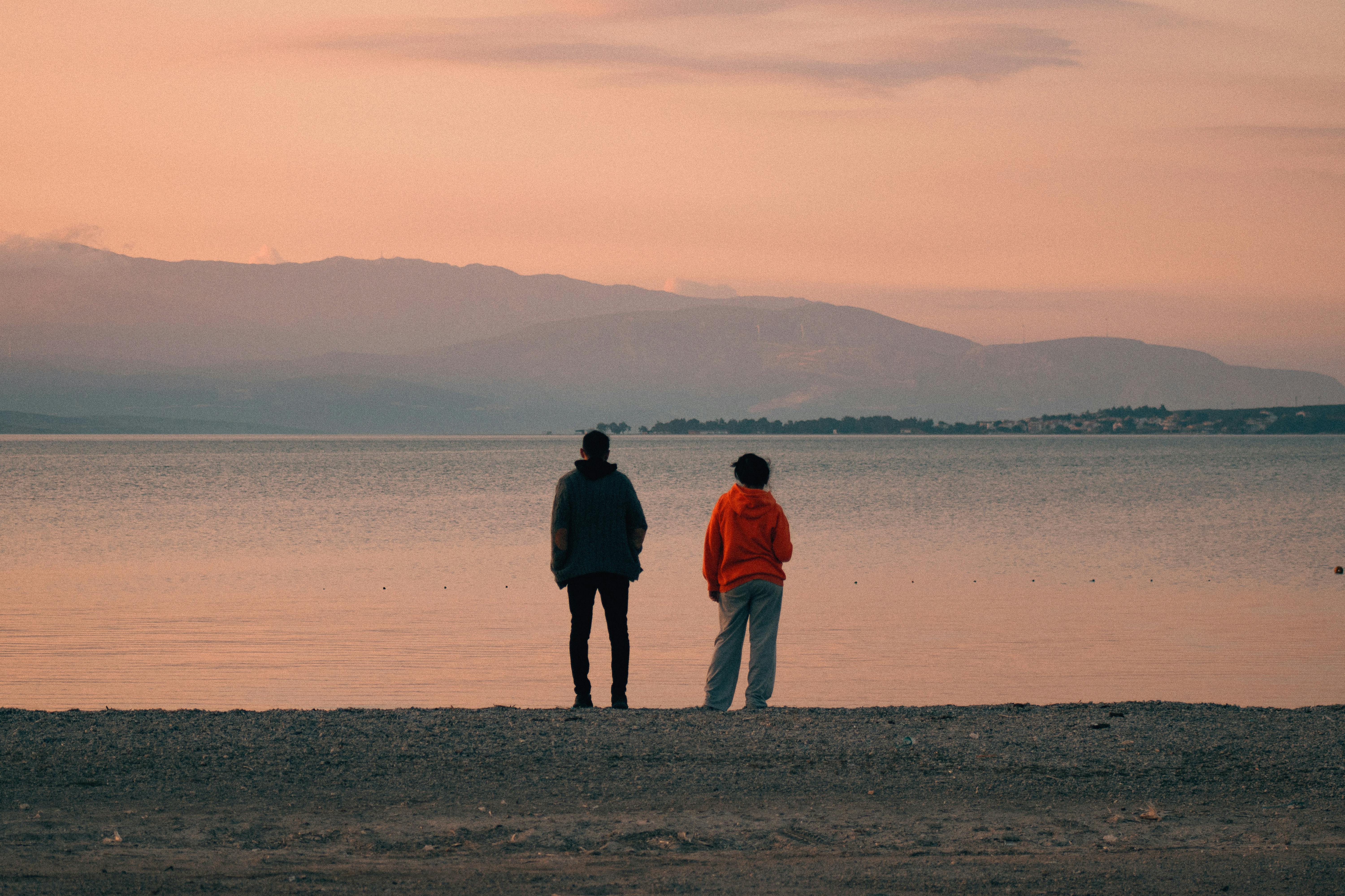 two people standing on the beach looking at the water