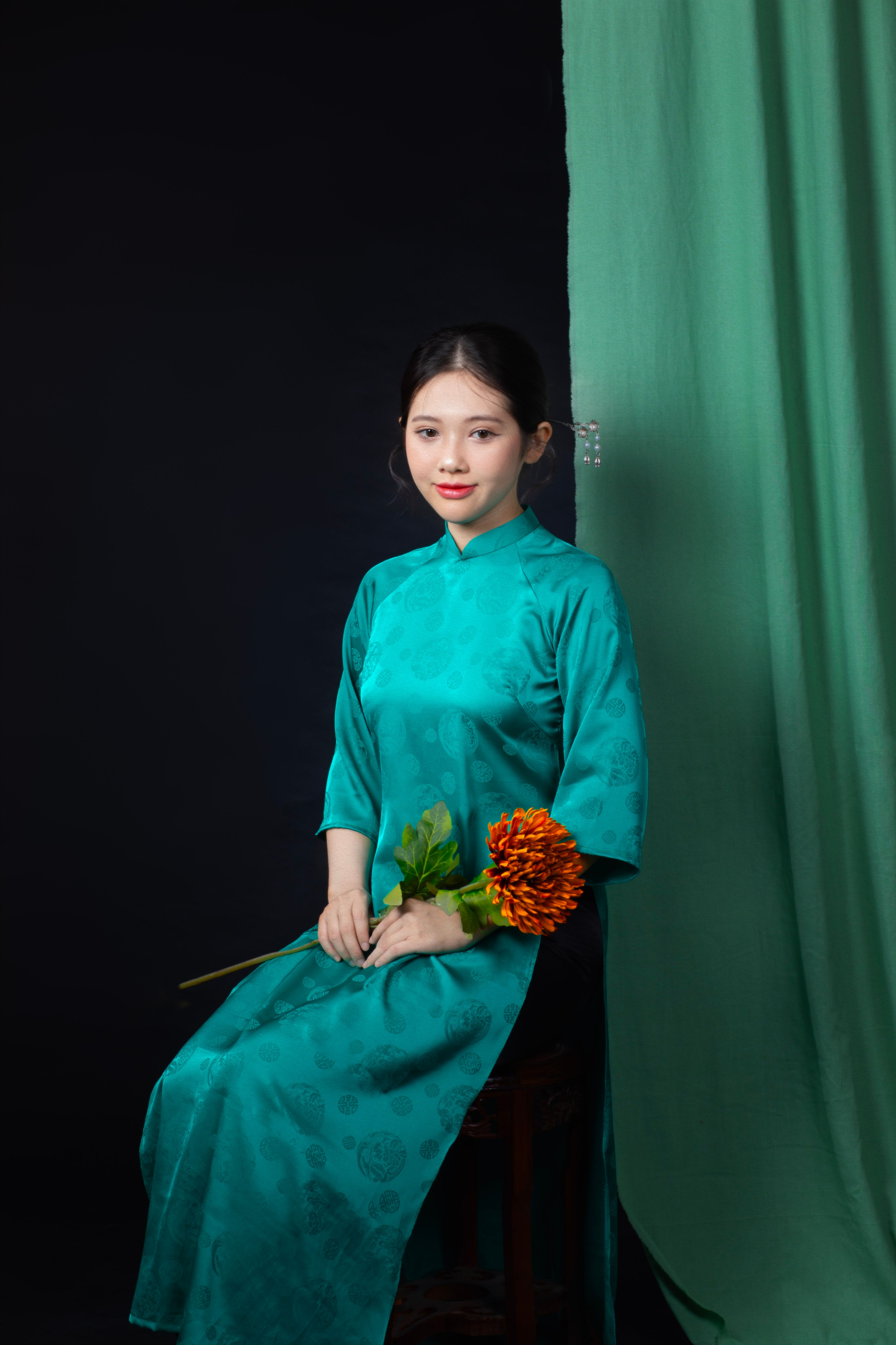 Young Vietnamese woman in a teal ao dai holding a flower, posing against a dark background.