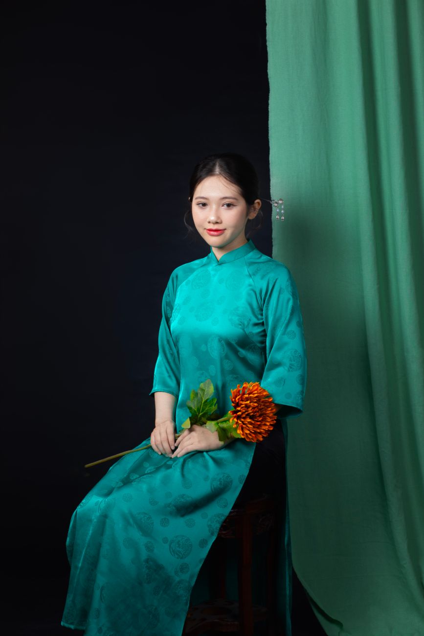 Young Vietnamese woman in a teal ao dai holding a flower, posing against a dark background.
