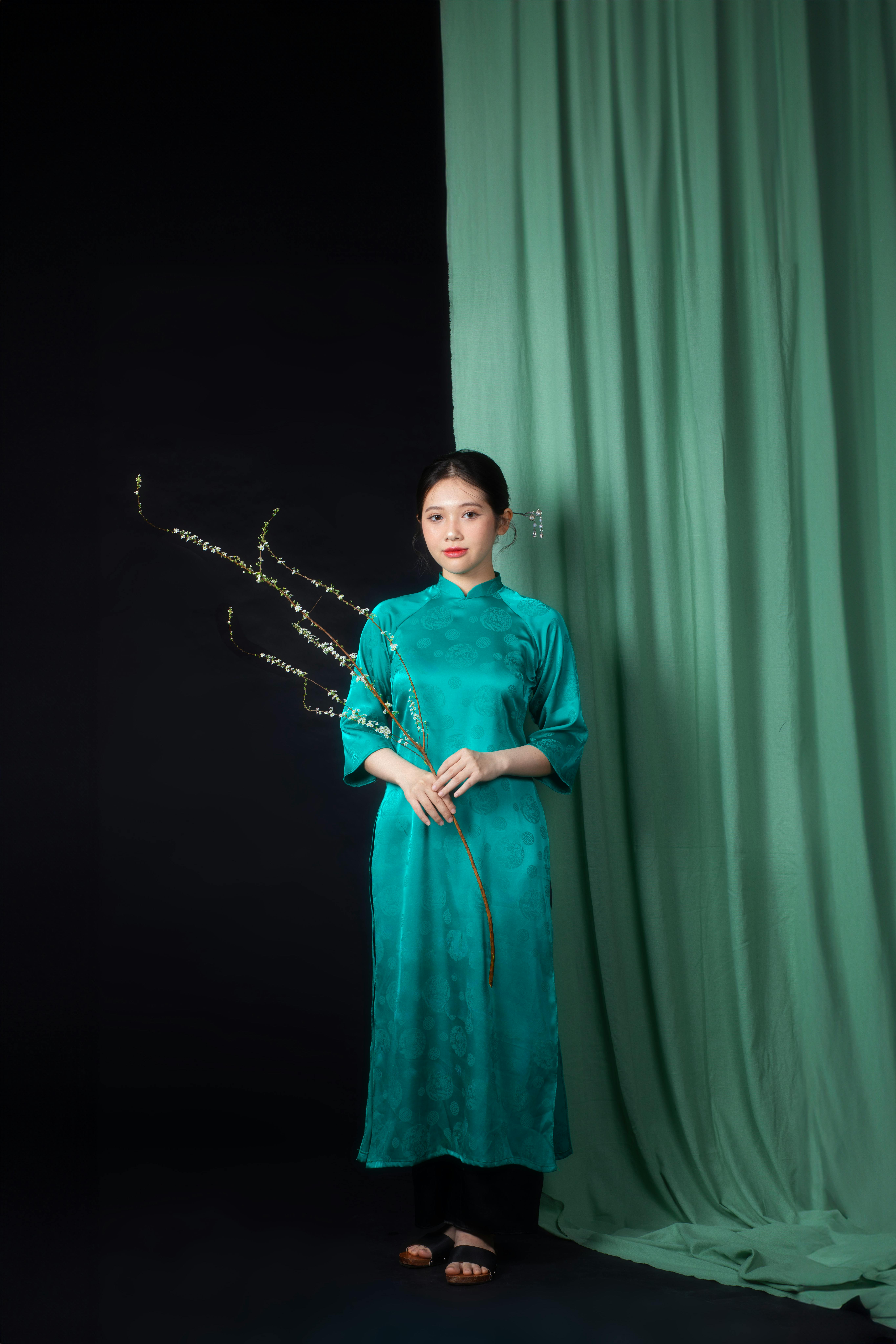 Elegant portrait of an Asian woman in traditional dress holding flowers against a green curtain backdrop.
