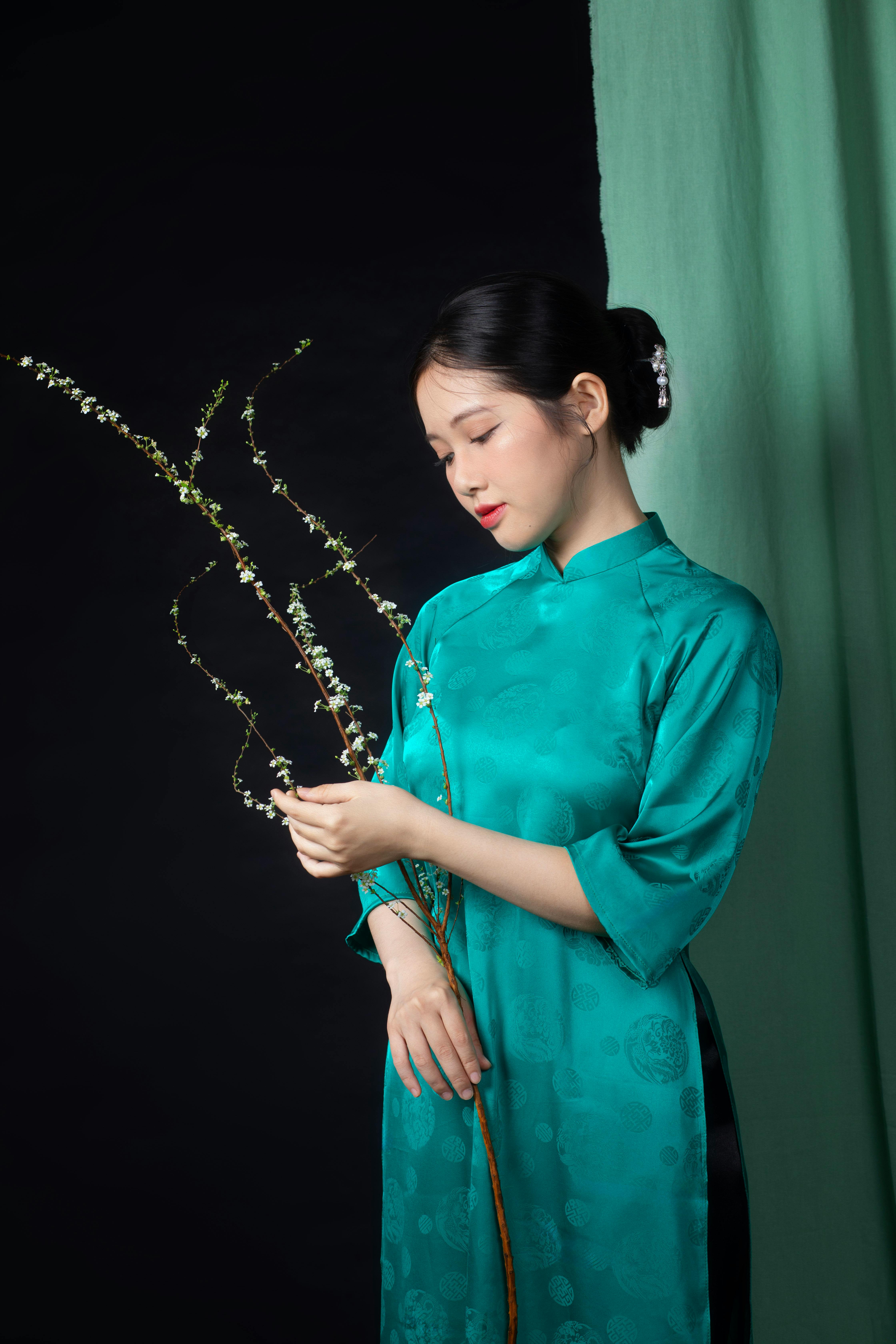 Serene portrait of a young woman in a turquoise ao dai holding twigs, studio setting.
