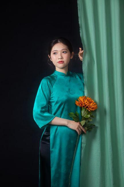 Portrait of a young Vietnamese woman in traditional ao dai, holding flowers and posing against a curtain.