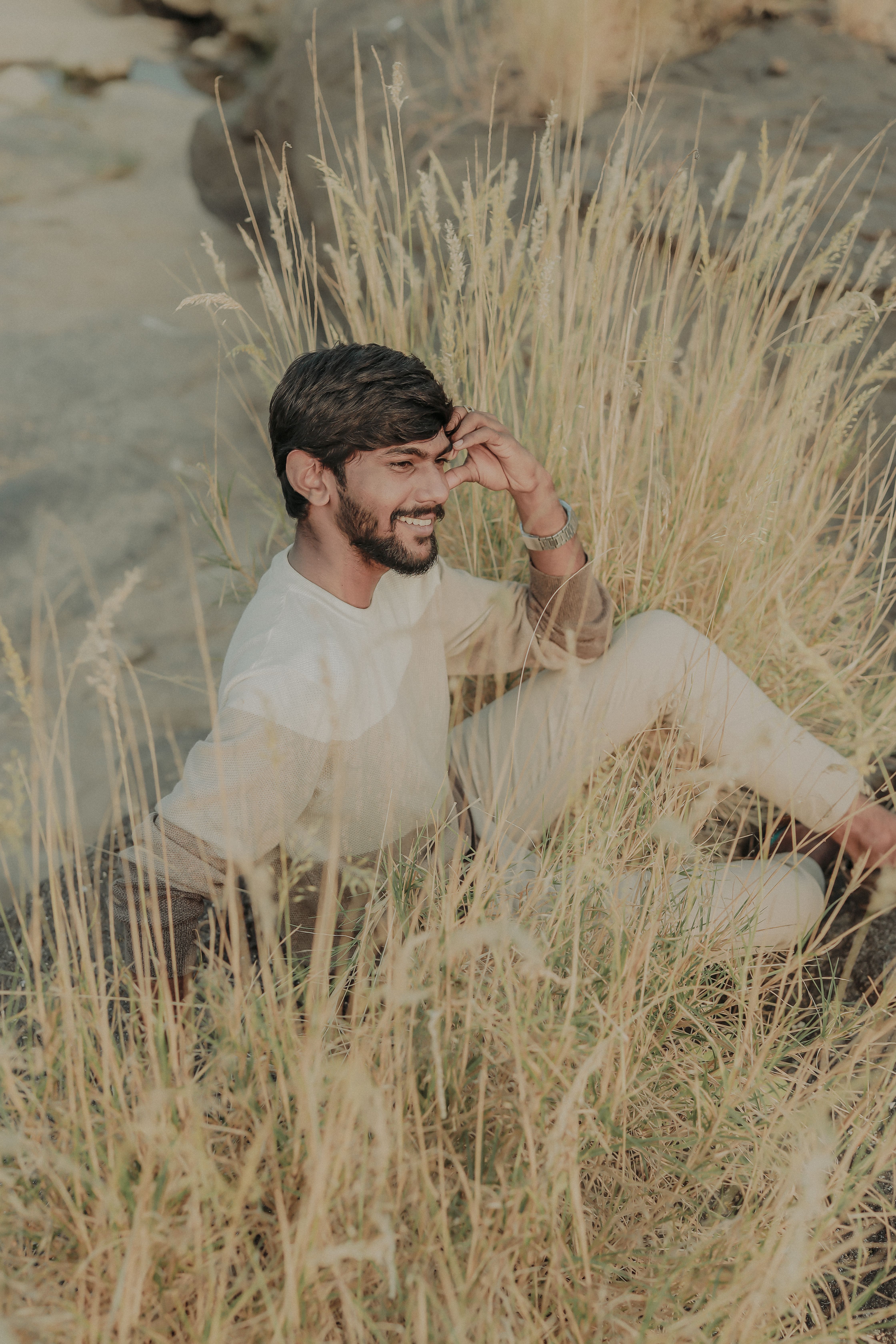 A relaxed man with a beard sits smiling among tall grass, enjoying a sunny day.