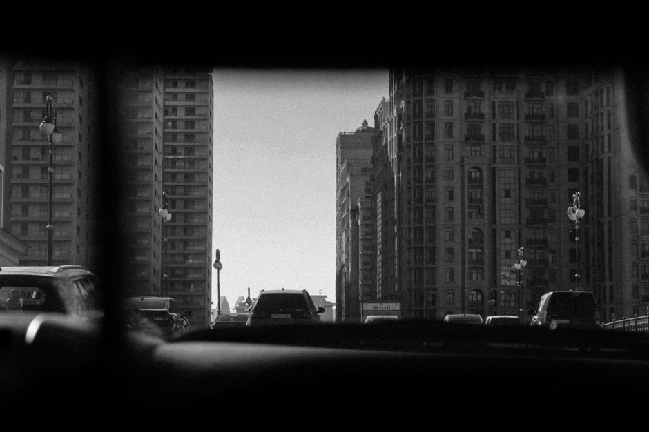 Black and white view of a city street through a car windshield, capturing urban architecture and vehicles.