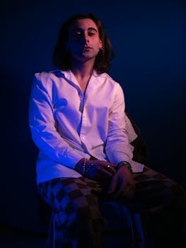 A man in a white shirt sits in dramatic blue lighting, exuding style.