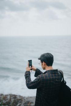 A young man stands by the sea, using his smartphone to capture the ocean horizon.