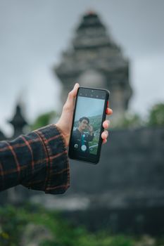 A person holding a smartphone, capturing a selfie with a temple in the blurred background.